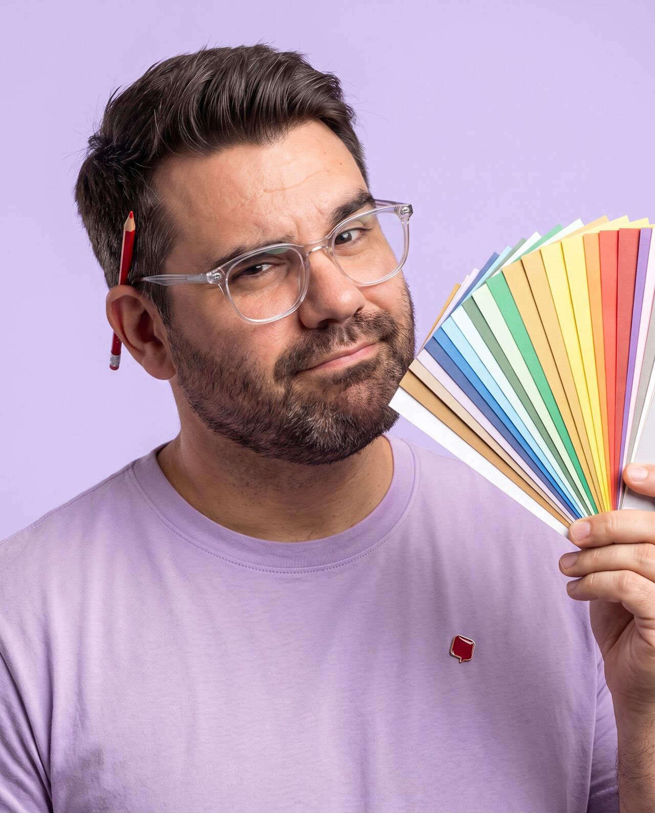 AI-generated portrait of Richard as a UI/UX designer in clear glasses and a lilac t-shirt, studying a fan of colour swatches with one eyebrow raised, a red pencil behind his ear