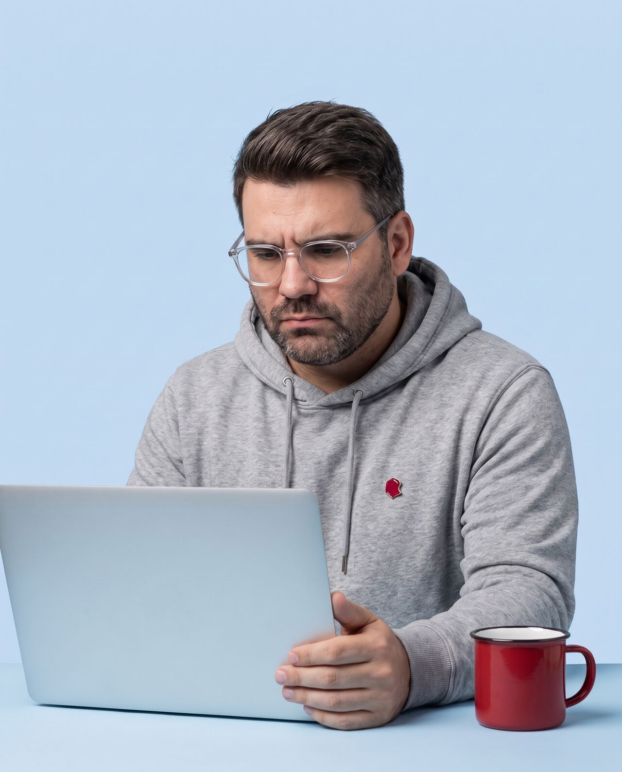AI-generated portrait of Richard as a developer in clear glasses and a grey hoodie, frowning in concentration at his laptop, a red enamel mug beside it, on a sky-blue backdrop